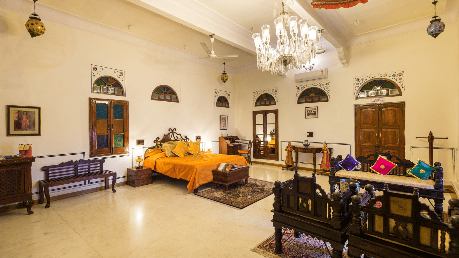 A well-lit bedroom at Fort Barli, featuring a vibrant orange bedspread, a crystal chandelier, and antique furniture, offers a spacious and elegant retreat.
