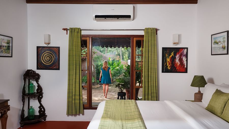 A shot of a room featuring a bed, a wooden corner rack, and a glass window from which a woman can be seen walking outside amidst the green surroundings - BluSalzz Villas - Muhamma Resort, Alleppey