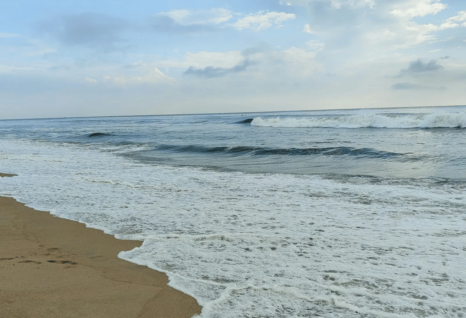 White sea foam and gentle ocean waves crashing onto a sandy beach under a light blue sky with scattered clouds.