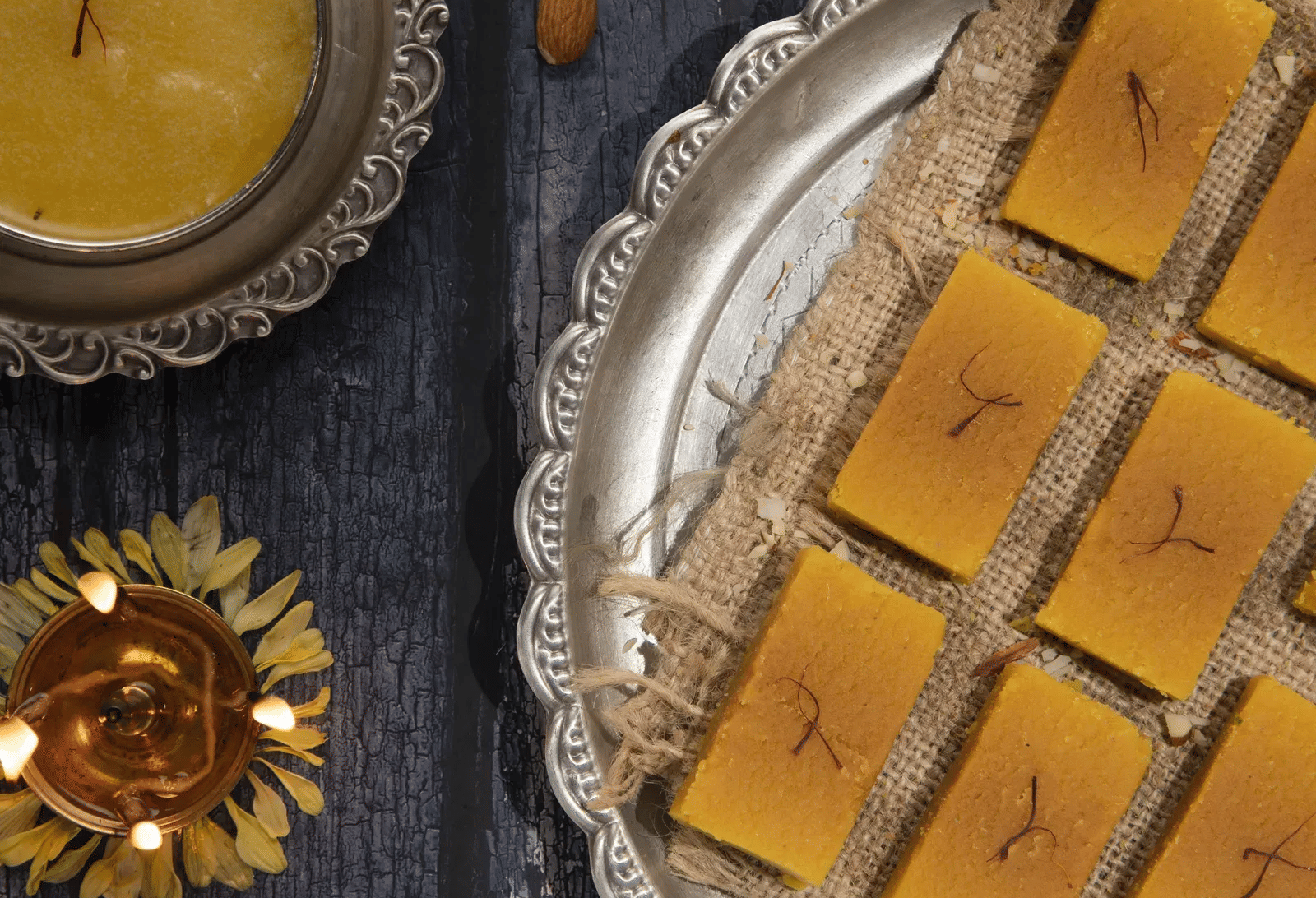 An overhead view of yellow Indian sweets arranged on a silver platter next to a small traditional clay diya lamp.