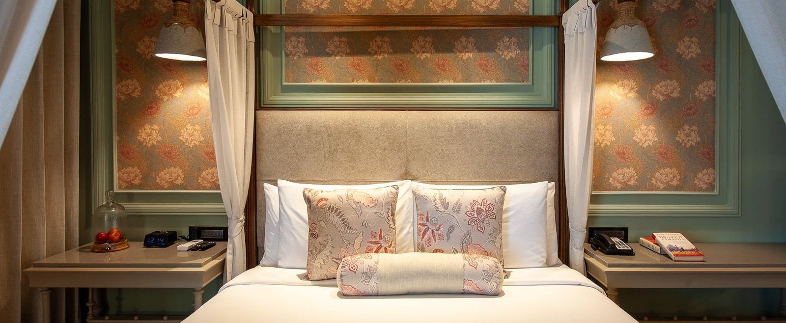 A close-up, front-facing view of the beautifully appointed four-poster bed in a suite at The Evren, Vagator. The bed is adorned with decorative pillows, and the background features elegant wallpaper and unique hanging lights.