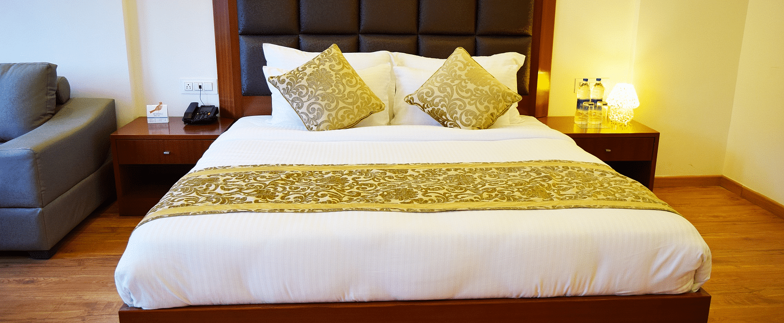 The image shows a hotel room with a large bed, two nightstands, and a couch in the background  | DLS Serenity Resort and Spa