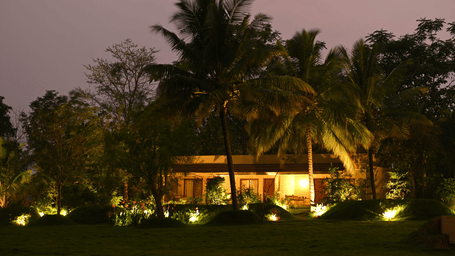 Fireflies Resort Kabini beautifully illuminated at twilight, surrounded by lush tropical greenery under a stunning purple-hued evening sky.
