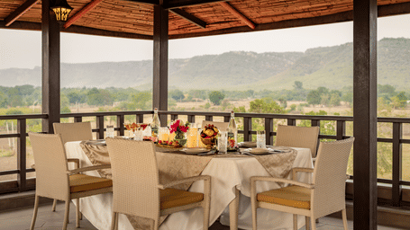 Covered outdoor dining terrace of Badal Mahal under a thatched roof with a set dining table overlooking distant green hills at AamaGhati Wildlife Resort.