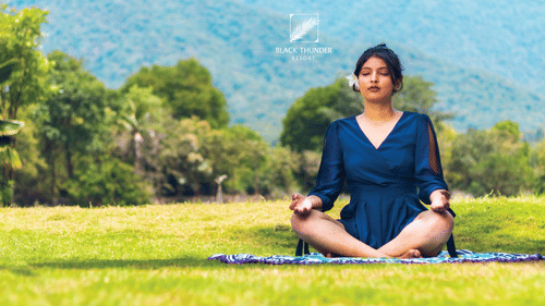 A woman meditating on a grassy field with mountains in the background, sitting cross-legged and focusing on mindfulness - Black Thunder, Coimbatore