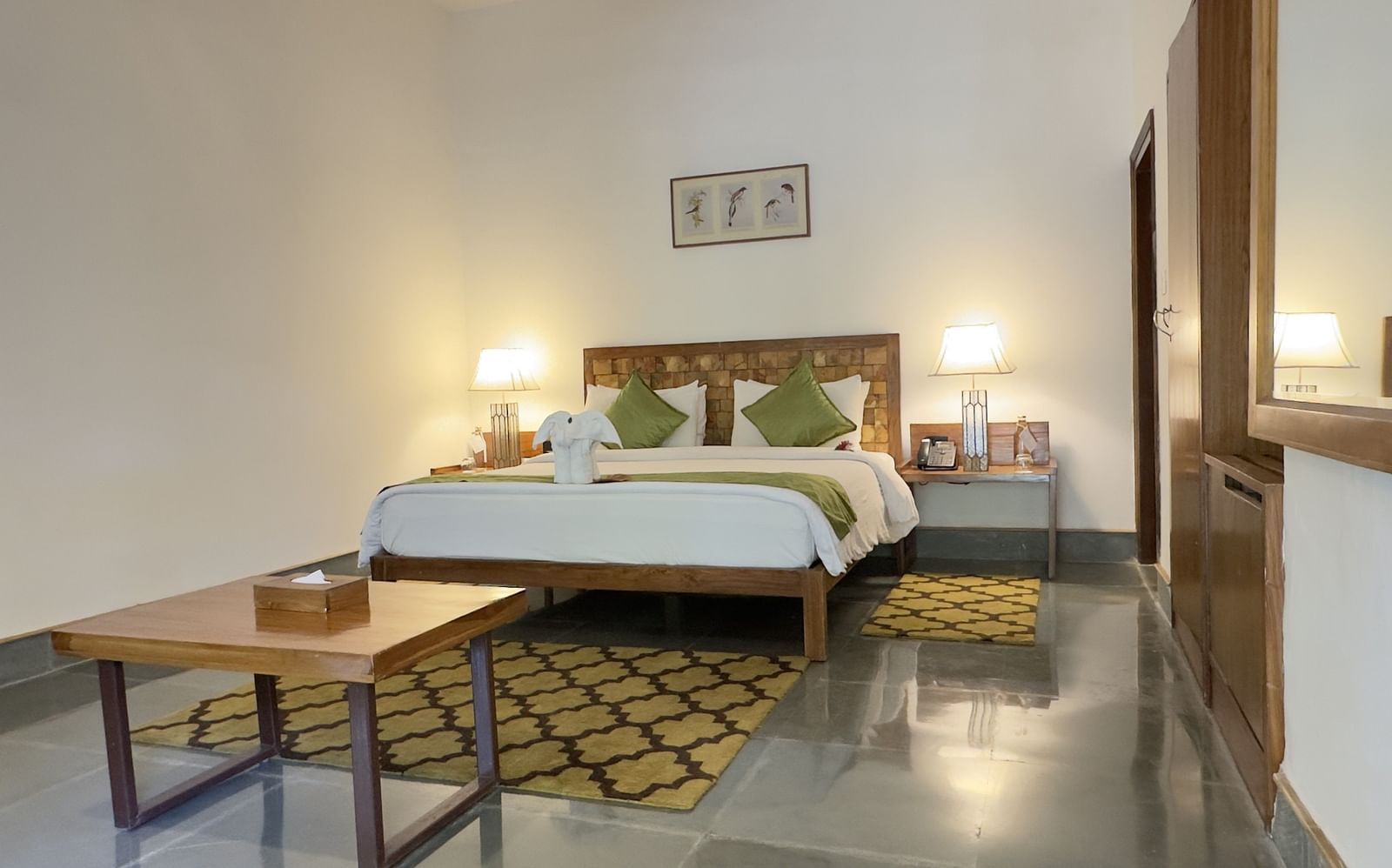 An image of a bedroom with double size bed, a centre table, bedside table on either side and lamps lit on top - Saraca Resort & Spa Corbett