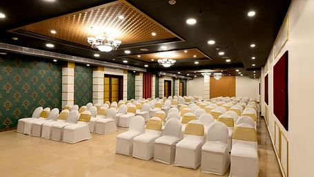A spacious banquet hall with chairs covered in white cloths and gold  covers. The hall has a modern design with dark wood accents and a crystal chandelier.