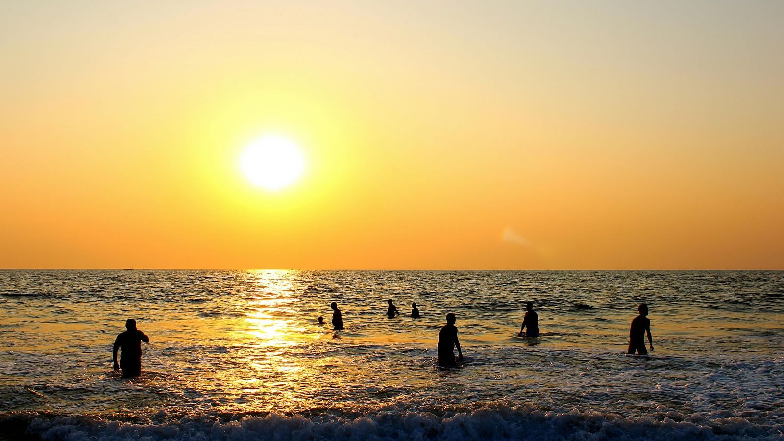 A beach featuring a beautiful sunset during the early evening with people bathing on the sea shore.