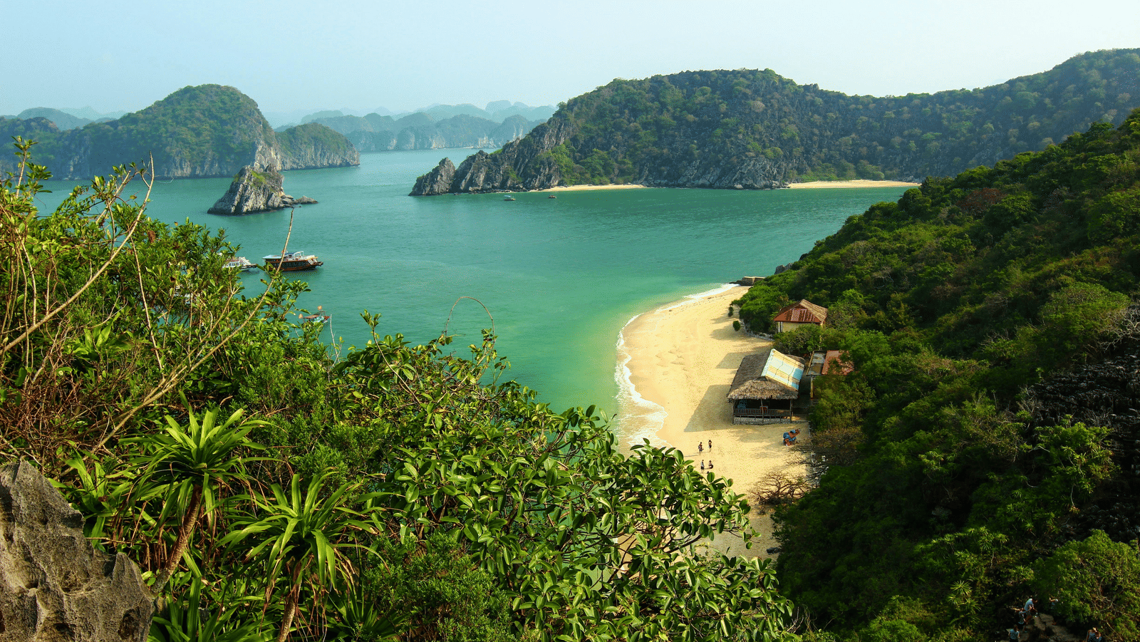  A high-angle view of a secluded beach with turquoise water, a few small huts, and several boats surrounded by lush green islands.