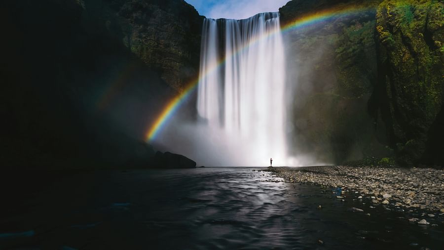 A person standing at the edge looking at the waterfalls with a rainbow in front of the falls