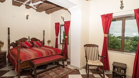 A spacious bedroom at Fort Barli, featuring a four-poster bed with red accents, checkered floors, and exposed wooden beams, offers a blend of rustic charm and elegance.