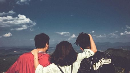 Three friends with their arms around each other stand on a mountain top looking out at a scenic view.
