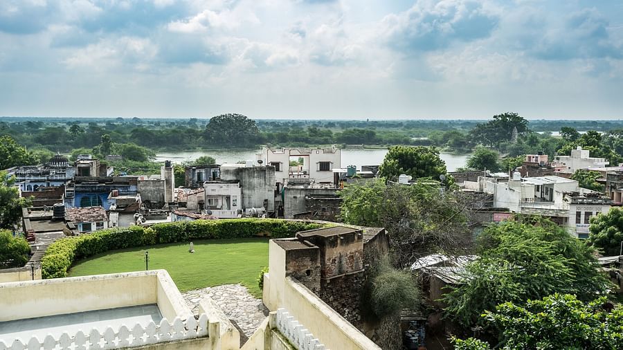 An elevated view from Fort Barli overlooks a townscape with a river and lush greenery, under a dramatic sky with clouds.