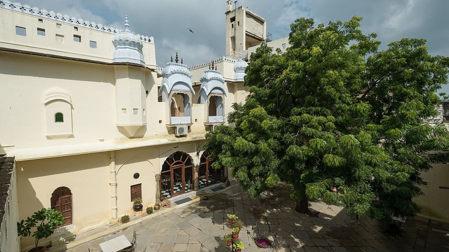 A courtyard at Fort Barli features a large tree, white buildings with decorative windows, and a distant tower against a cloudy sky.