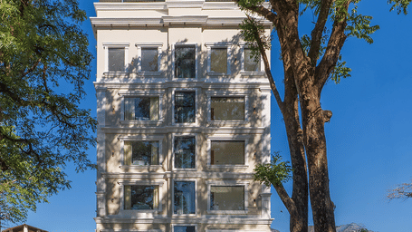 A tall, narrow white building of Perfectstayz Premium Aurevva, featuring windows behind a paved entrance area, flanked by large green trees under a clear blue sky.