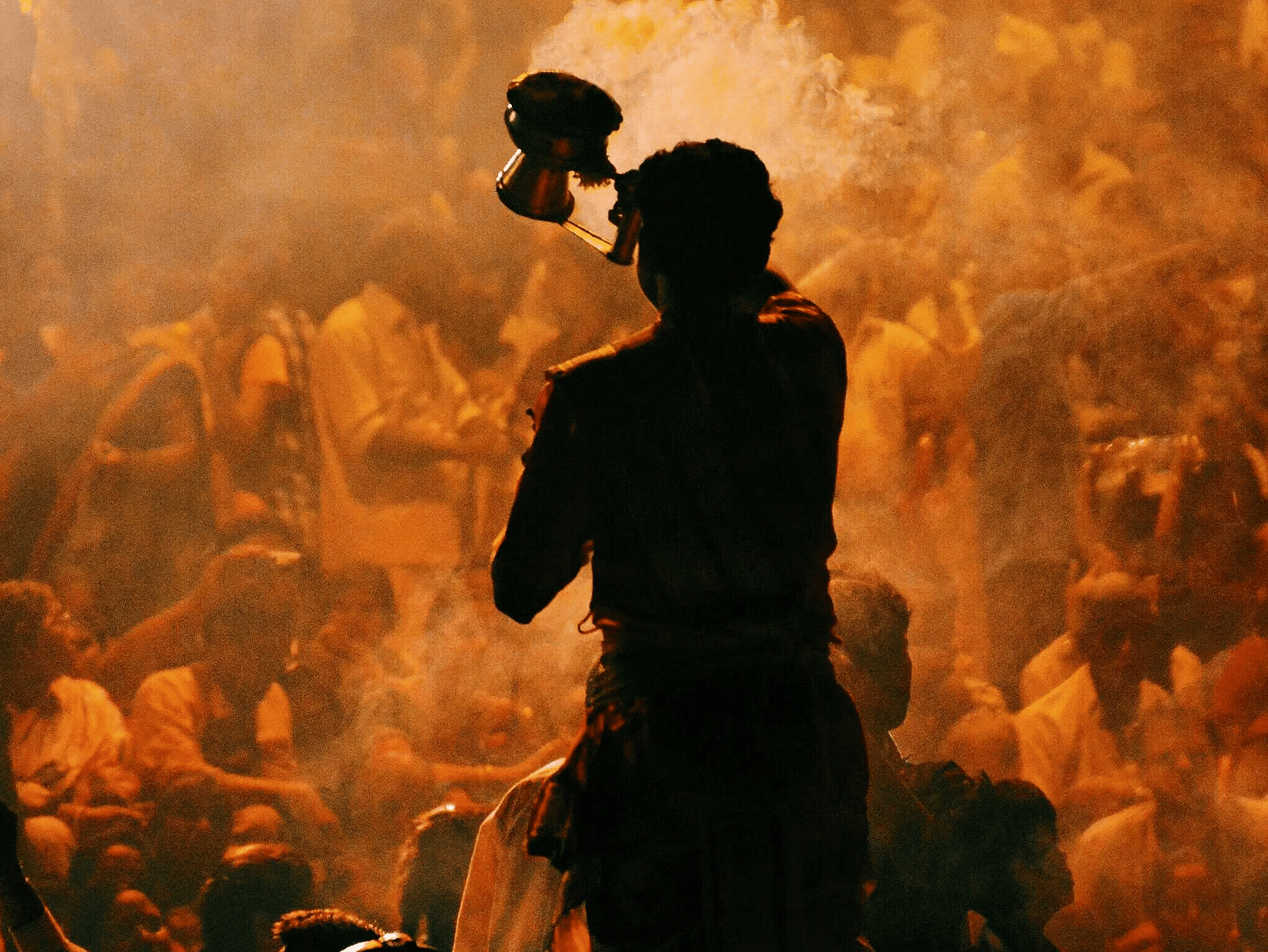 A silhouetted figure holds up a small object amidst a crowd bathed in warm, hazy light.