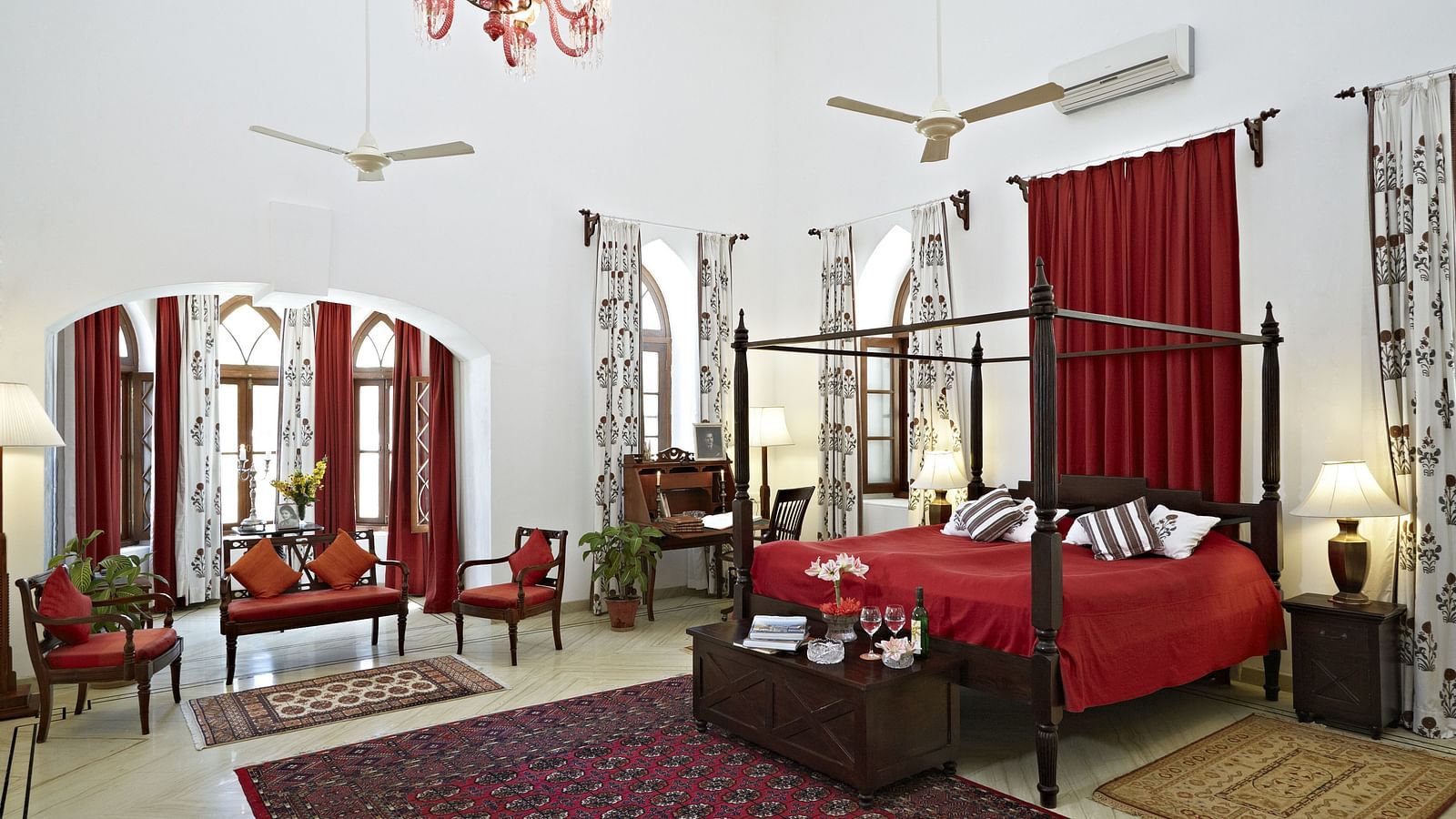 A spacious room featuring a four-poster bed with linens, seating arrangements, decorative rugs, and arched windows with draped curtains- Shahpura Bagh.