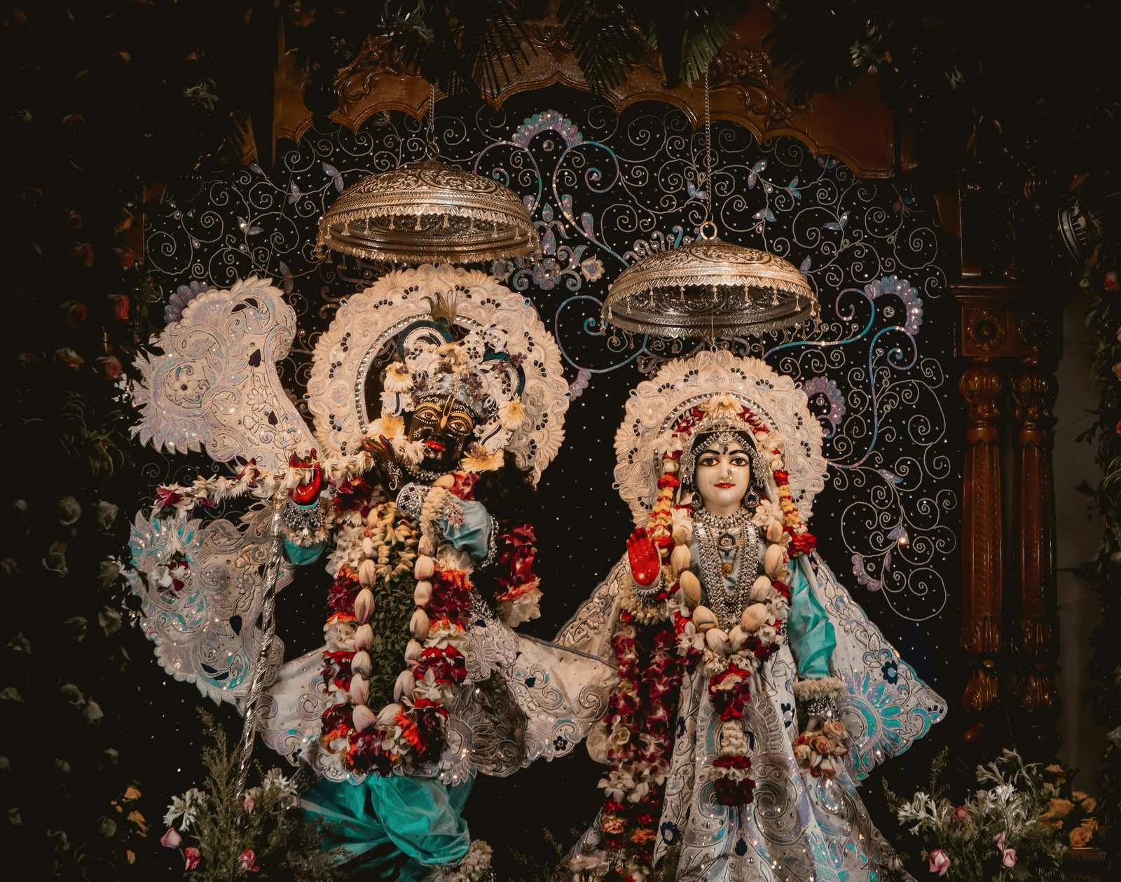 Close-up of 2 Hindu idols or deities adorned with flowers and silver ornamentation, against a dark, ornate background.