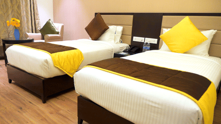 Premium Twin Room at ASR Parklane, Hosur, featuring twin beds, a nightstand with 2 water bottles, and a seating area.