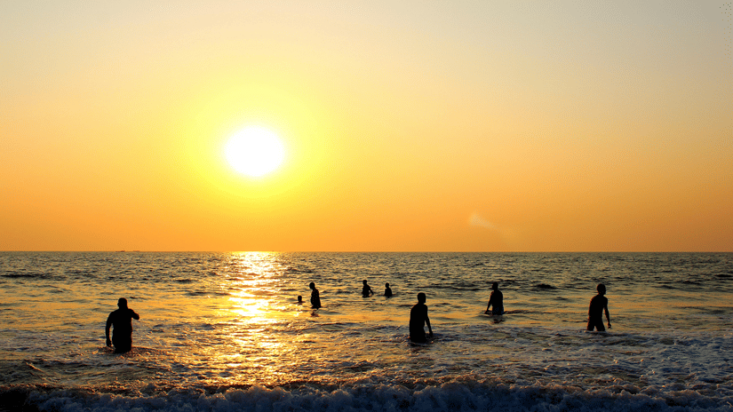 The silhouettes of people swimming in the ocean at sunset, with a bright orange sun reflecting on the gentle waves.
