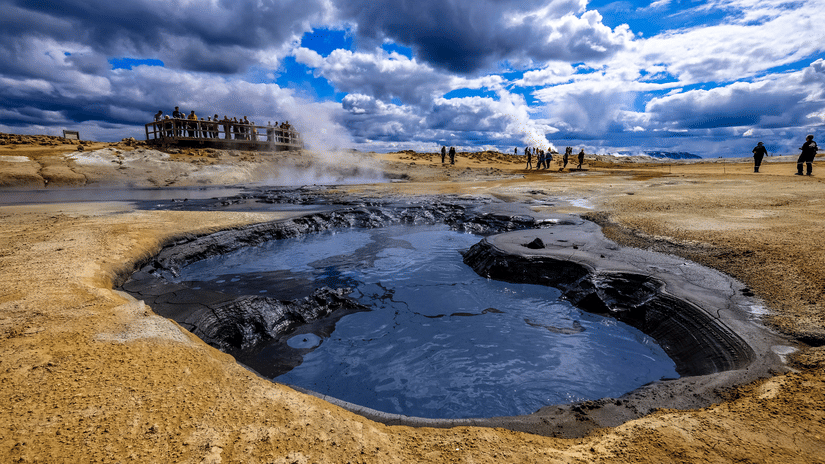 A geothermal hot spring surrounded by rocky ground under a  cloudy sky.