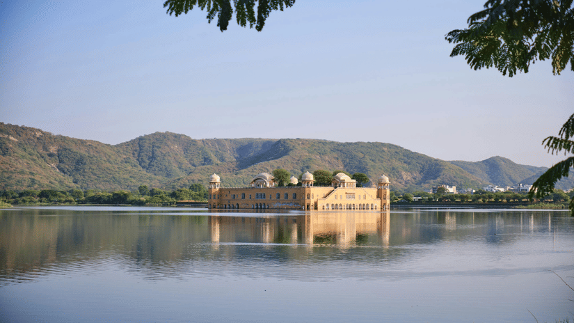 The Jal Mahal palace sits serenely in the middle of Man Sagar Lake, framed by rolling green hills and a blue sky.