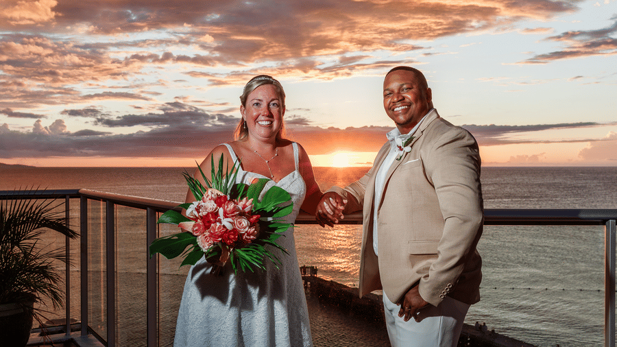 A bride and groom smiling on a balcony overlooking the ocean at sunset, with the bride holding a vibrant bouquet and warm golden light in the background.