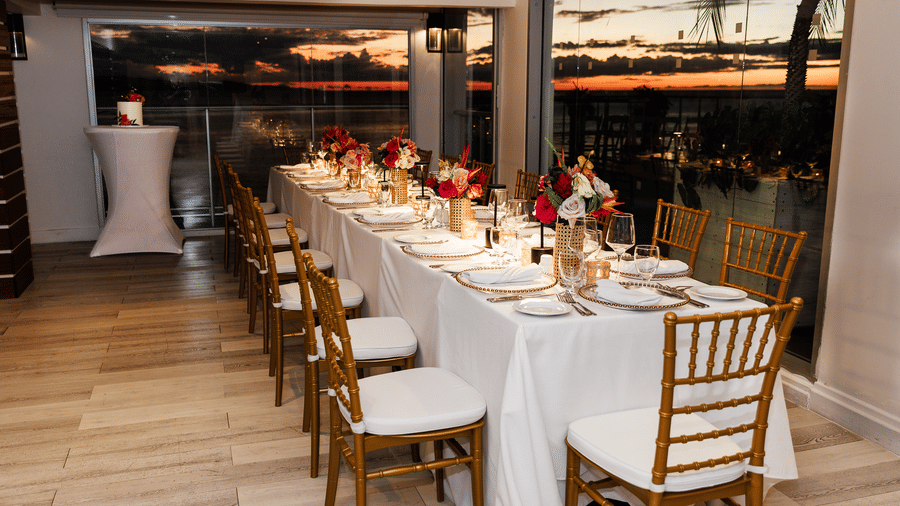 A long dining table elegantly set with white linens, gold chairs, and floral centrepieces inside a glass-walled venue, overlooking a scenic sunset view.
