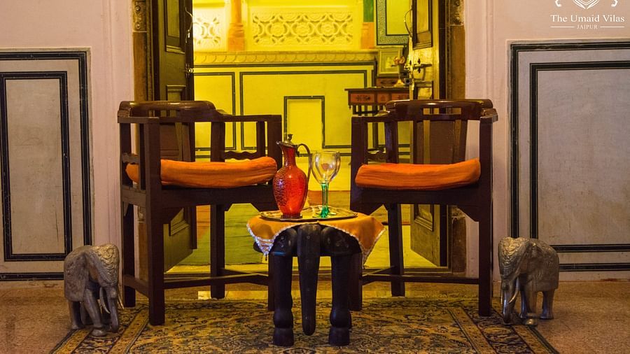 A seating area at Umaid Vilas Jaipur with two wooden chairs, orange cushions, a small table with a red bottle and glasses, and elephant sculptures on either side.