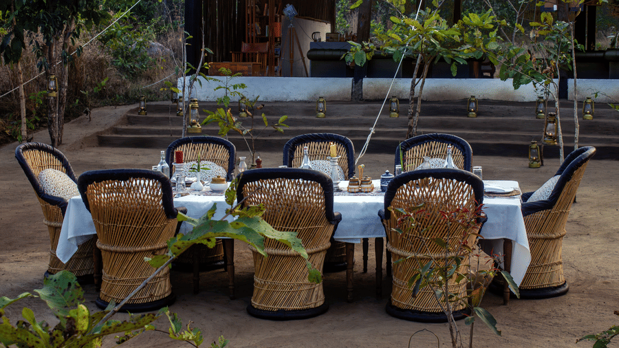 Wicker dining chairs around a linen-covered table, viewed through foliage, prepared for a meal near a stone structure - Umaria Safari Camp