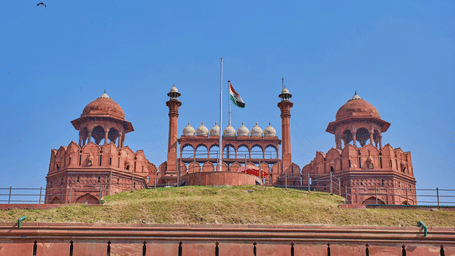 Side view of the Red Fort in New Delhi featuring its red sandstone walls and domes under a clear blue sky with green lawns in the foreground.