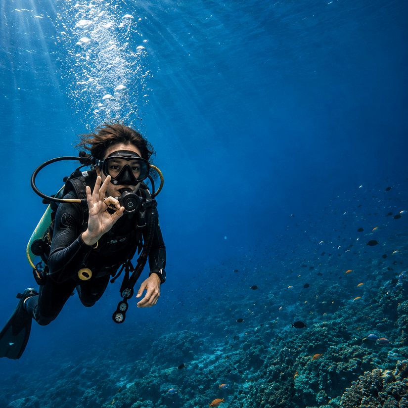 A scuba diver swimming underwater above a coral reef in clear blue ocean water.