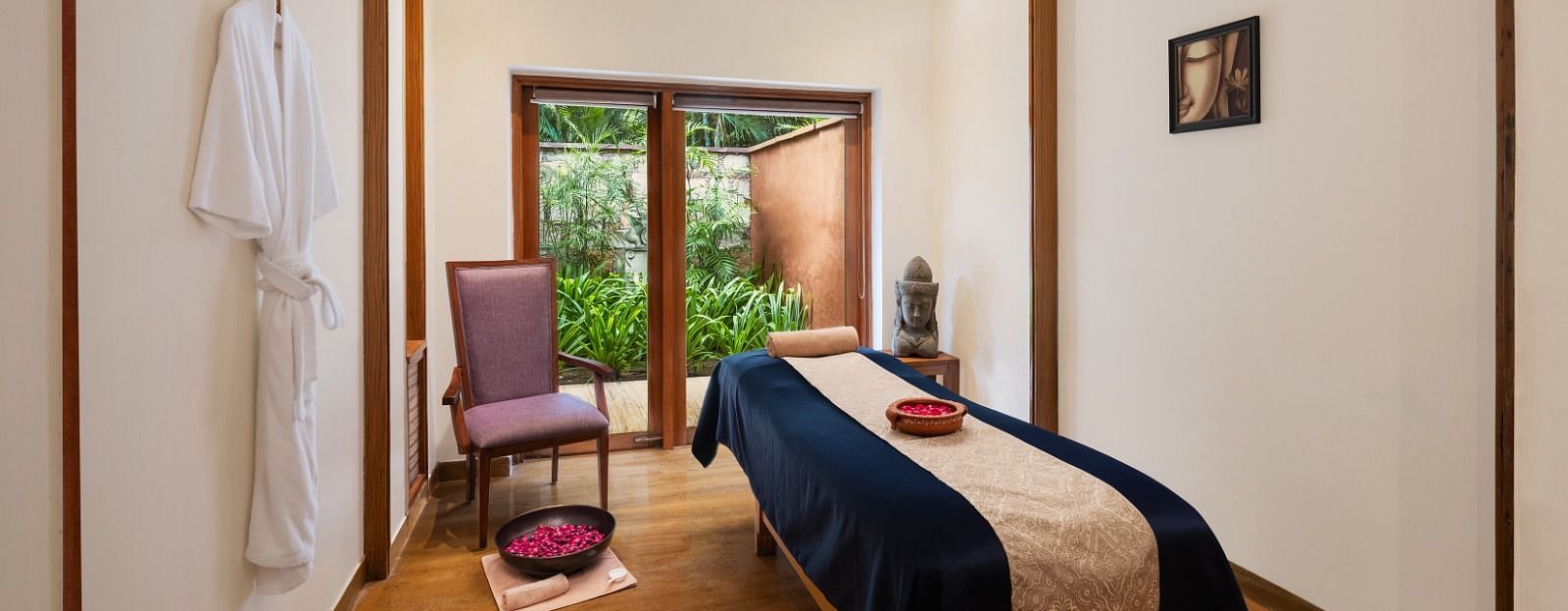 A serene spa room with a massage table, soft lighting, a chair, a robe hanging on the wall, and a window overlooking greenery - Ananta Spa and Resort, Pushkar.