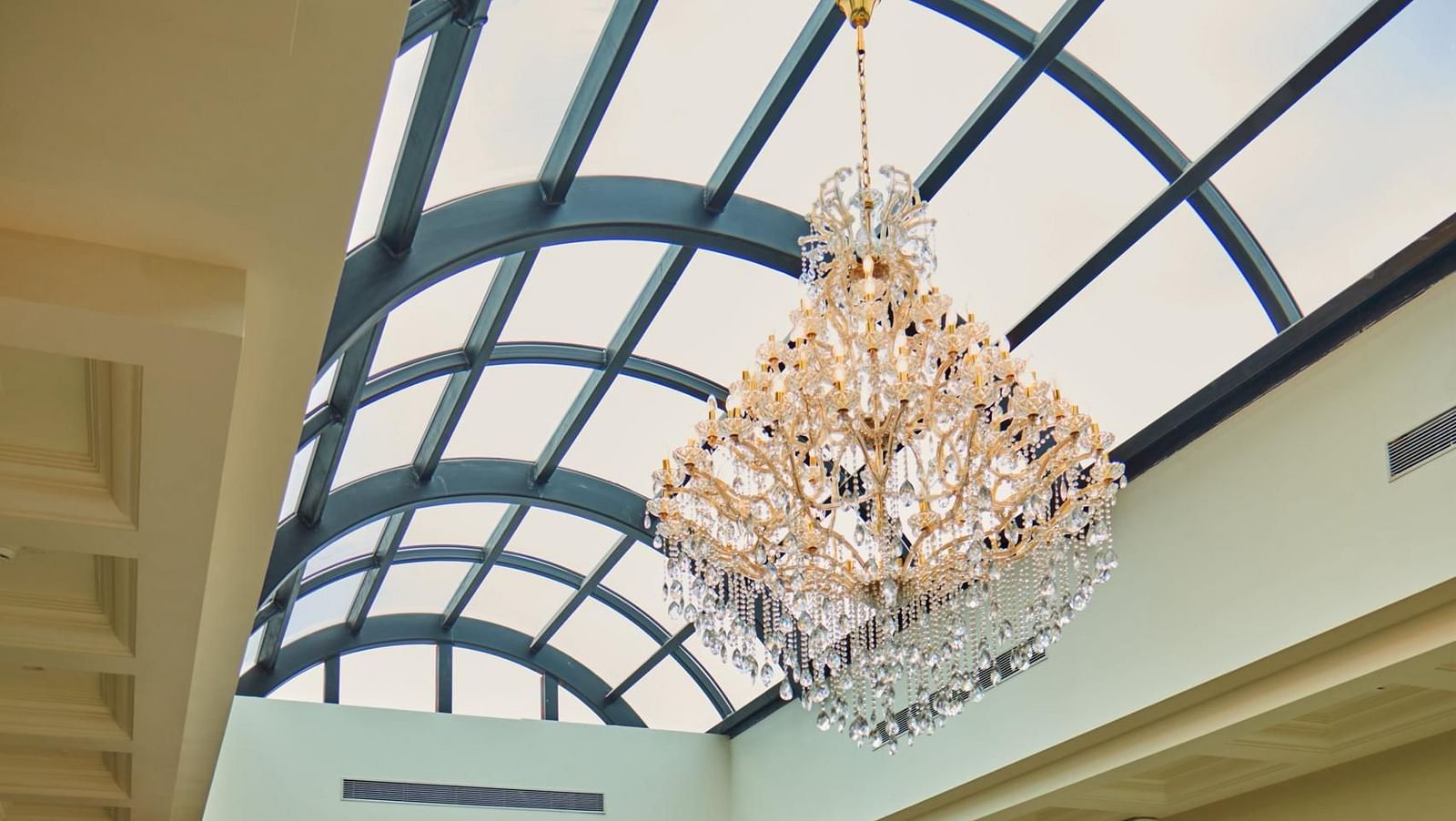 A spacious, elegant indoor Meli-Melo with a high arched glass ceiling, vibrant seating, a central fountain, chandeliers, large windows, and potted plants creating a bright, inviting atmosphere