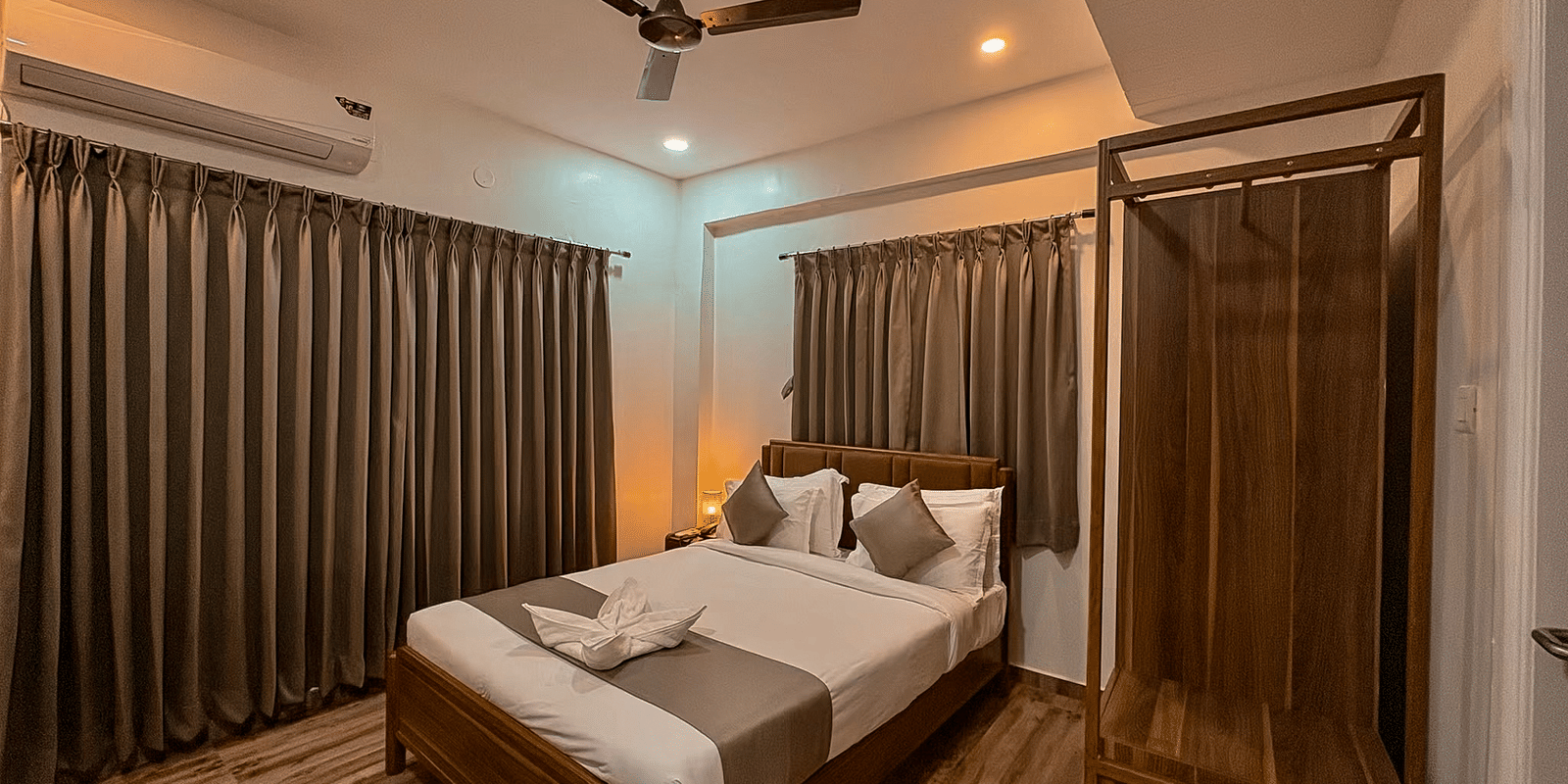 Wide veiw of a bedroom in the Coral 4BHK of Kove Smart Stays featuring wooden flooring, plush bedding, a wardrobe unit, and closed curtains above the headboard of the bed, and one whole wall of the room.