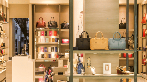 Boutique window display with rows of leather handbags in black, tan, and blue, along with high-heeled shoes and wallets on lit shelves.
