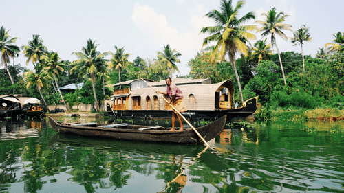A person poles a small boat through the calm backwaters, with a traditional houseboat and lush green palm trees lining the waterway.