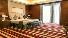 Modern guest room at The Boma Nairobi with king bed, wood panelled walls, bedside lamps, large window with curtains, and colourful striped carpet.
