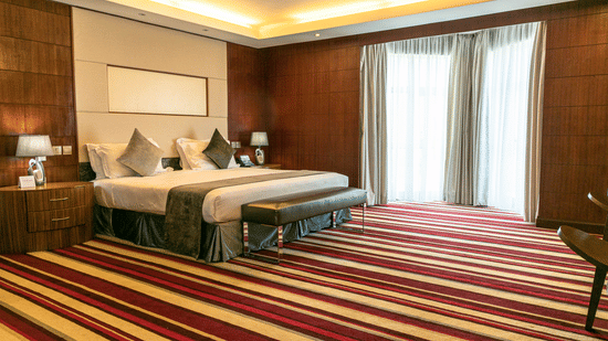 Modern guest room at The Boma Nairobi with king bed, wood panelled walls, bedside lamps, large window with curtains, and colourful striped carpet.