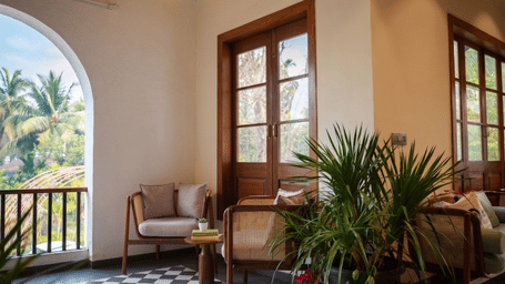 Armchairs surround a small wooden table near a potted plant, tall glass doors, a chequered tiled floor at Bougainvillea, Alibaug.