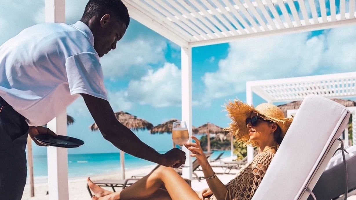A waiter hands a drink to a woman relaxing on a lounge chair under a shaded cabana on a sunny beach.