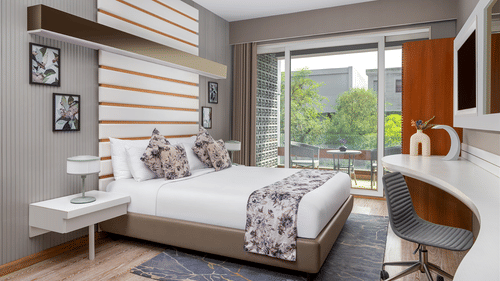 A modern bedroom at The Atara, Gurugram featuring a neatly made bed with a floral runner, a stylish white desk with an armchair, and a large window offering a refreshing view of lush greenery