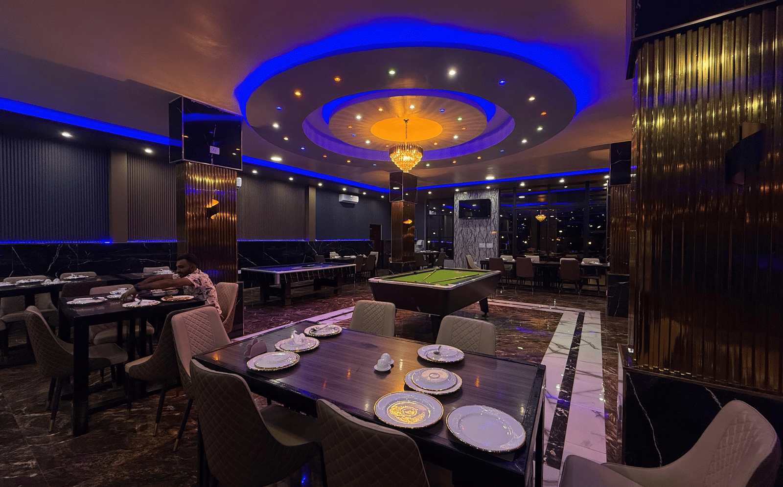 A dark, luxurious recreation room with a pool table, set tables, and illuminated by a central chandelier and blue neon ceiling lights at Luffu Club.