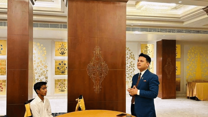 A speaker addresses an audience inside a hall at Clarks Group of Hotels. A table stands next to the presenter.