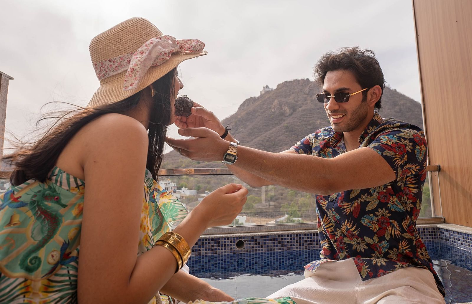 Two people sitting outdoors. The person on the left is wearing a hat and has long dark hair. The person on the right is wearing sunglasses and a patterned shirt.