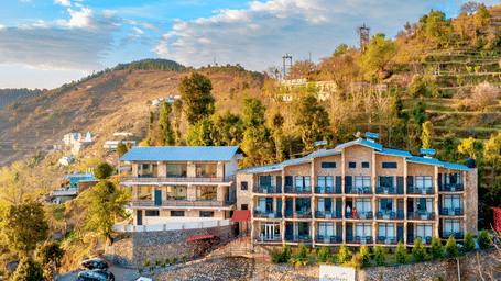 An aerial view of the multi-storey resort building situated on a green hillside under a blue sky at Himalayas Resort By The Lake Hill, Mukteshwar.