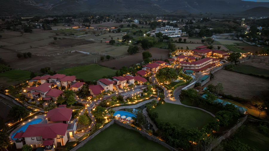 Aerial view of the sprawling layout of AamaGhati Wildlife Resort at dusk featuring warm illuminated villas, swimming pools, pathway lighting, and a vast green lawn against distant mountains.
