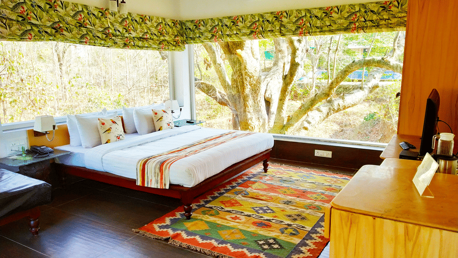 Tree house at The Riverwood Forest Retreat, Pench featuring a double bed, a bedside table, a TV atop a cabinet, an A/C unit, and windows with curtains drawn providing a view of outside.
