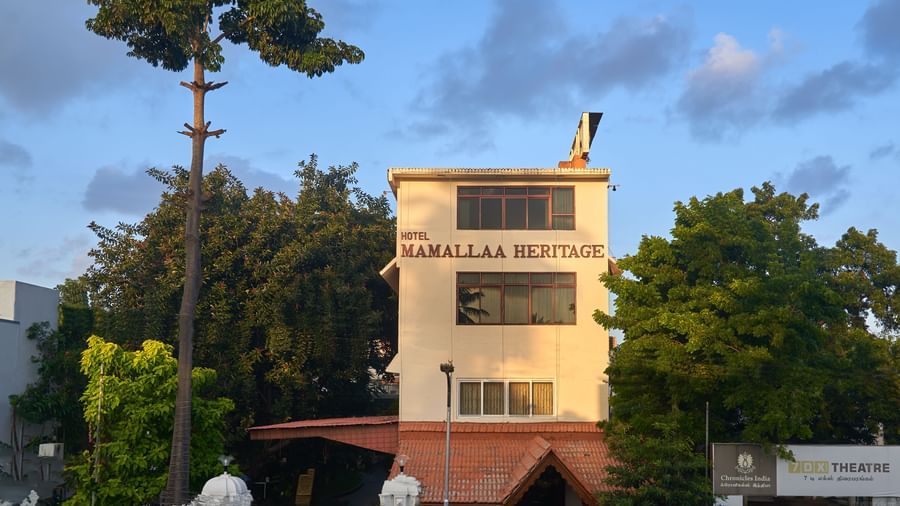 The multi-story exterior of Hotel Mamallaa Heritage, a light-coloured building with a prominent sign, surrounded by green trees under a blue sky.