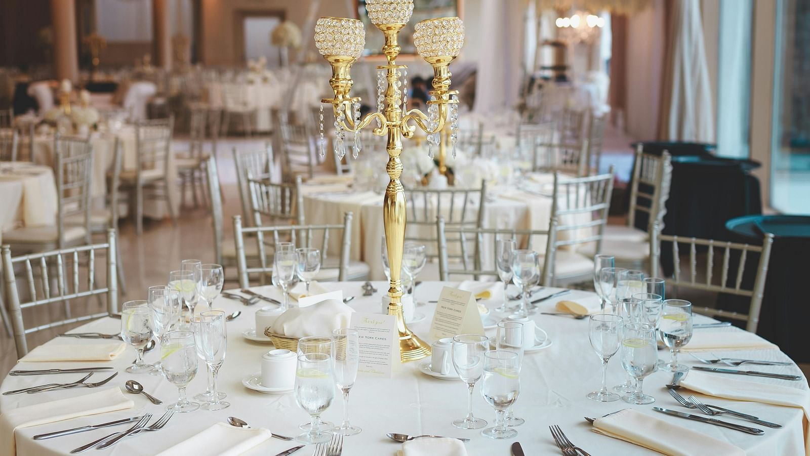 A formally set round table, featuring a white tablecloth, elegant place settings with silverware, glassware, and napkins, and a tall, gold candelabra as a centrepiece, all set within a large room with additional tables and chairs in the background.
